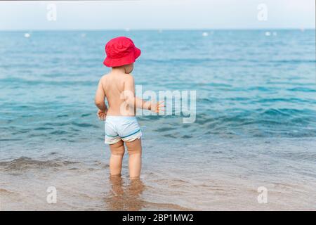 Cute adorable little blond caucasian serious pensive toddler boy in red panama walking through blue clear water waves of tropical sea or ocean beach