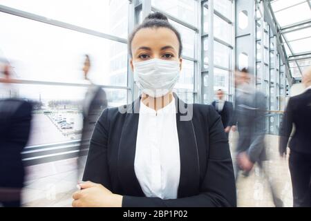 Beautiful african business woman in suit wearing surgical protect mask standing in a crowd of walking people. Concept for prevent for coronavirus outb Stock Photo