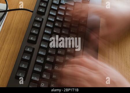 Blurred hands due to fast typing on a black keyboard on a wooden desk in a home office Stock Photo