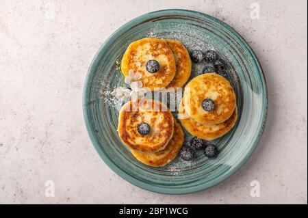 Traditional russian cheesecake with blueberries and powdered sugar on a ceramic plate on a light background. Top view. Copy space for text Stock Photo