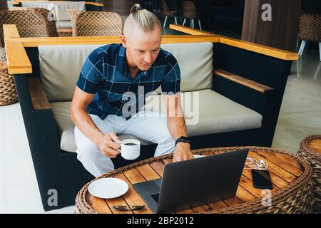 A young guy sits in a cafe, holds a cup of coffee, checks the message on a laptop, and writes a response letter. Morning Post. The gaze is directed to Stock Photo
