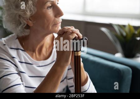 Thoughtful elder disabled woman sitting on sofa with wooden stick. Stock Photo