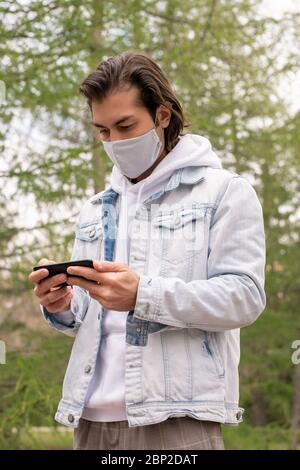 Busy young man in casualwear and protective mask watching video in smartphone while standing in public park during pandemic period Stock Photo