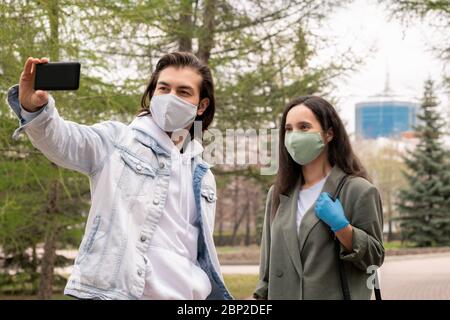 Young man and his girlfriend in casualwear and protective masks making selfie in public parkwhile enjoying walk on warm summer day Stock Photo