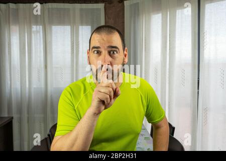 Bearded man with short hair in green shirt making silence symbol in living room at home. Tranquility concept Stock Photo
