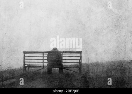 A mental health concept of mysterious figure back to camera,  sitting on a bench alone. With a grunge, textured edit. Stock Photo