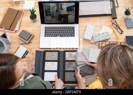 Two young female creative designers sitting by table in front of laptop and discussing samples of marble tiles, linoleum and home interior photo Stock Photo