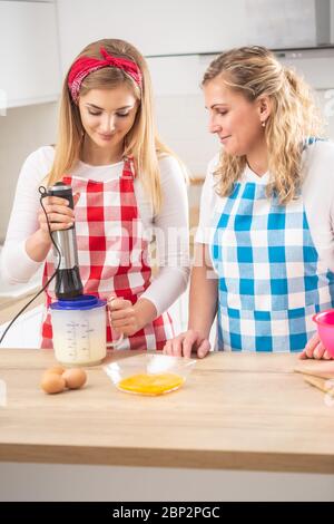 Mother helping daughter in teh kitchen, with the girl mixing eggs with a hand blender. Stock Photo