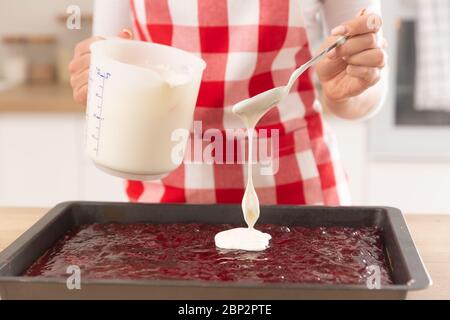 Detail of a woman pouring whisked egg whites from a spoon on a jam topping of a cake in a baking tray. Stock Photo
