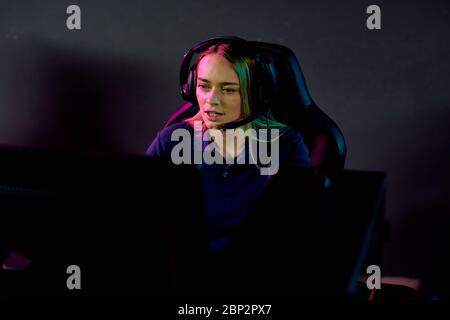 Young serious blond female e-sport gamer in headset looking at computer screen while sitting in dark club in front of monitor Stock Photo
