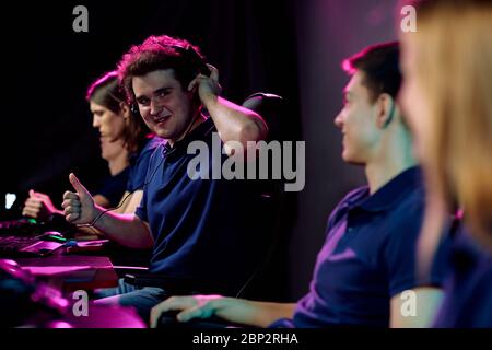 Young successful e-sport gamer showing thumb up and looking at one of friends with smile while playing cyber game in front of monitor Stock Photo