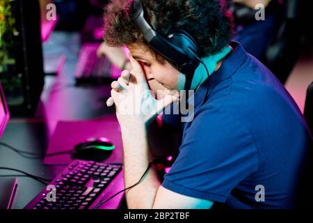 Tense or worried young man in headset touching face while slightly bending over keyboard in front of computer monitor Stock Photo