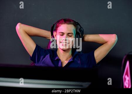 Young smiling relaxed male video gamer in headphones enjoying break between e-sport games while keeping his hands behind head Stock Photo
