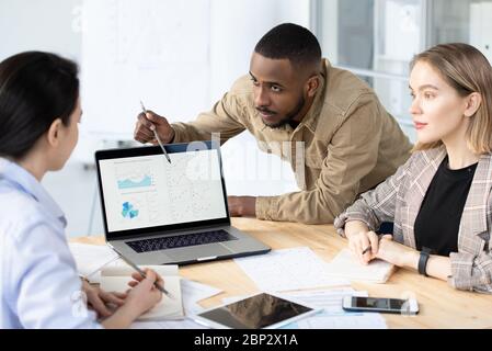 Young confident banker or broker bending over desk while analyzing financial data on laptop display and listening to colleague Stock Photo