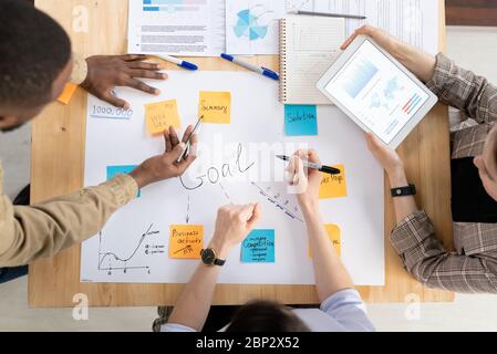 Top view of three intercultural managers or analysts discussing business goals written on notepapers stuck to big paper at startup meeting Stock Photo