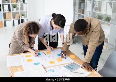 Three intercultural colleagues with highlighters bending over table with papers and writing down ideas on notepapers while brainstorming Stock Photo