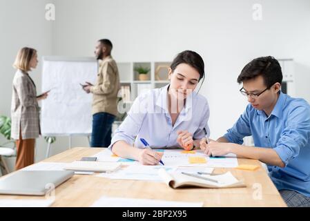 Two young intercultural brokers sitting by table in office and analyzing data or making working notes on background of their colleagues talking Stock Photo