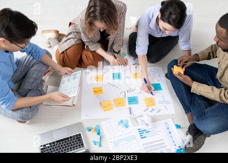 Four intercultural startuppers in casualwear sitting on the floor of office and getting ready for conference while discussing new ideas Stock Photo
