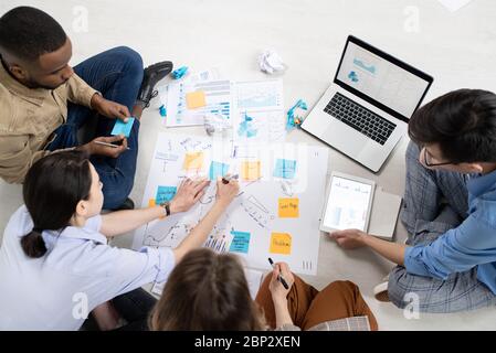 Group of intercultural brokers analyzing financial information and documents in front of laptop while sitting on the floor around papers Stock Photo