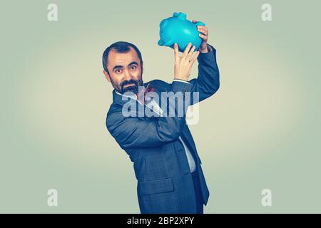 Adult displeased bearded man in elegant suit with red bow tie holding overturned piggy bank, showing its emptiness. Mixed race bearded model isolated Stock Photo