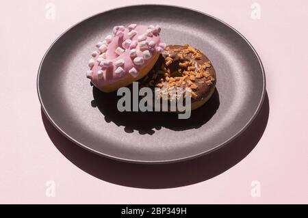 Two donats, one decorated with white marshmallows and another chocolate one with caramel-flavored curls on the the dark grey ceramic plate on pink Stock Photo