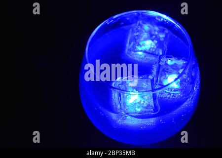 Blue bright glowing ice cubes that float in a delicious frosted white cold drink are located in a cognac glass on a black frosted background. Stock Photo
