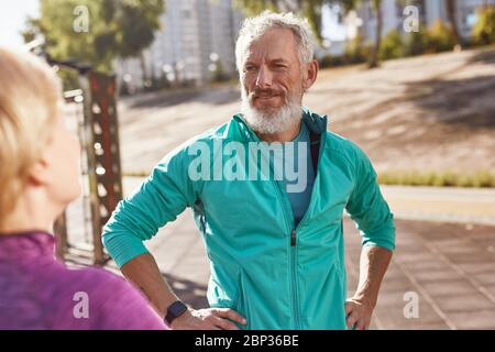 Athletic mature bearded man in sportswear talking with his wife while exercising together in the early morning, senior couple doing sport outdoors Stock Photo
