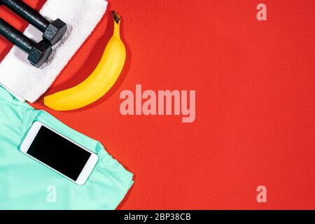 Set of sports attributes. Weights, a towel, a sports shirt, a mobile phone and a banana on a red mat for sport. Stock Photo