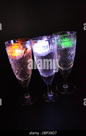 Crystal champagne glasses with carvings in the form of snowflakes are located on a black matte background. Bright multicolor doidic glowing ice cubes Stock Photo