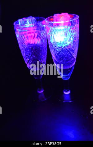 Crystal champagne glasses with carvings in the form of snowflakes are located on a black matte background. Bright multicolor doidic glowing ice cubes Stock Photo