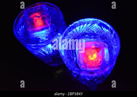Crystal champagne glasses with carvings in the form of snowflakes are located on a black matte background. Bright multicolor doidic glowing ice cubes Stock Photo
