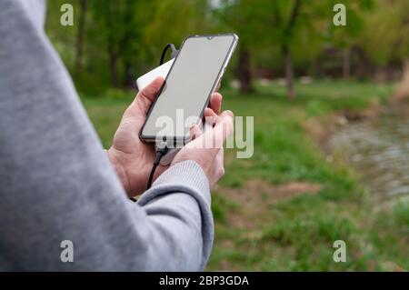 Man uses smartphone while charging from the power bank on the river's bank. Modern technology concept. Selective focus Stock Photo