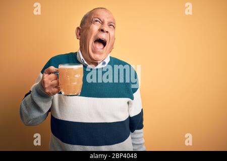 Senior handsome man drinking jar of beer standing over isolated yellow background angry and mad screaming frustrated and furious, shouting with anger. Stock Photo