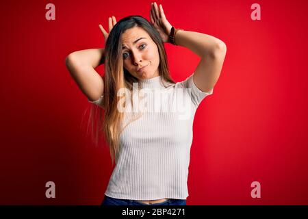 Beautiful blonde woman with blue eyes wearing casual white t-shirt over red background Doing bunny ears gesture with hands palms looking cynical and s Stock Photo
