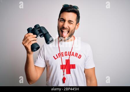 Young lifeguard man with beard wearing t-shirt with red cross and sunglasses using whistle sticking tongue out happy with funny expression. Emotion co Stock Photo