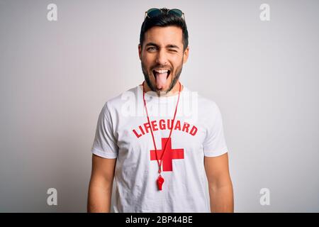 Young handsome lifeguard man with beard wearing t-shirt with red cross and whistle sticking tongue out happy with funny expression. Emotion concept. Stock Photo