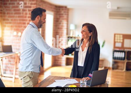 Two middle age business workers smiling happy and confident. Working together with smile on face using laptop doing handsake at the office Stock Photo