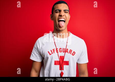 Young handsome african american lifeguard man wearing t-shirt with red cross and whistle sticking tongue out happy with funny expression. Emotion conc Stock Photo