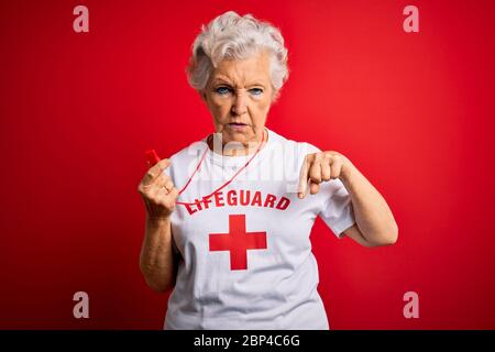 Senior beautiful grey-haired lifeguard woman wearing t-shirt with red cross using whistle Pointing down looking sad and upset, indicating direction wi Stock Photo