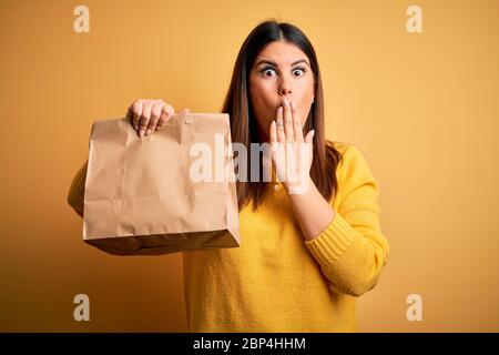 Young beautiful woman holding take away paper bag from delivery over yellow background cover mouth with hand shocked with shame for mistake, expressio Stock Photo