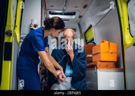 An injured, shocked man sitting with an oxygen mask in an ambulance, woman in a medical uniform is checking his pulse Stock Photo