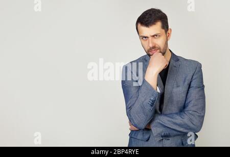 The man is looking thoughtfully at the camera. Confident young man with a beard in a stylish jacket, holds his hand on his chin, raised eyebrow thinks about something makes a plan, ponders a cool idea Stock Photo
