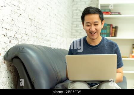 Young smiling Asian man in casual clothing on sofa working on laptop computer. Male freelancer working from home on notebook. Work from home concept Stock Photo