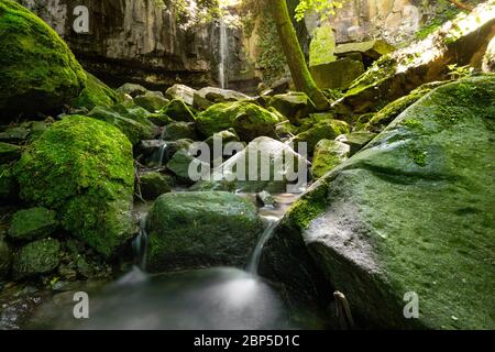 Wide angle view of a small mountain waterfall, with a creek running between moss-covered rocks and boulders and into a small pond Stock Photo