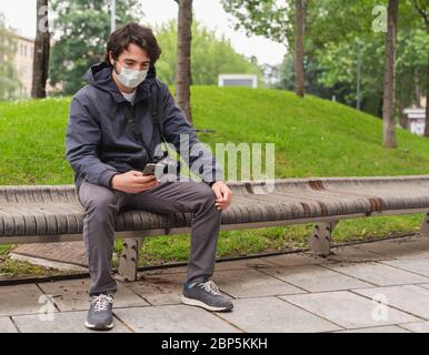 Young tourist with surgical mask sitting on a bench in a city in outbreak of Covid19, photographer with health care mask in urban exploration typing m Stock Photo