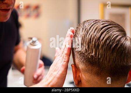Male hairdresser styles the hair of a young man with his hand and hairspray in his barbershop Stock Photo