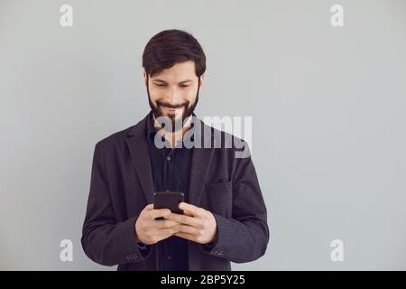 Positive guy in a jacket is typing text in a mobile smiling with on a gray background. Stock Photo