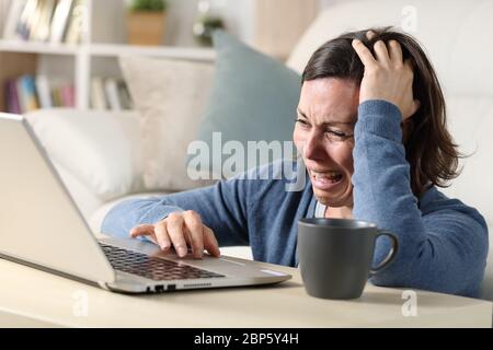 Sad adult woman crying reading bad news on laptop sitting on the floor on a coffee table at home Stock Photo