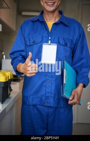 Smiling young repairman in blue uniform holding clipboard and showing thumbs-up Stock Photo