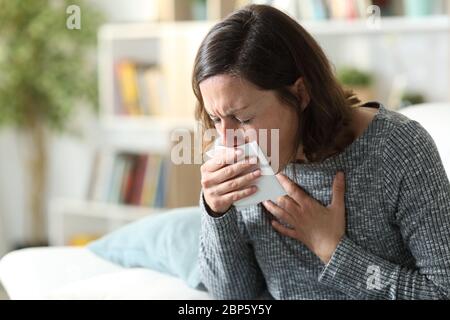 Sick adult woman coughing covering mouth with tissue sitting on a couch at home Stock Photo